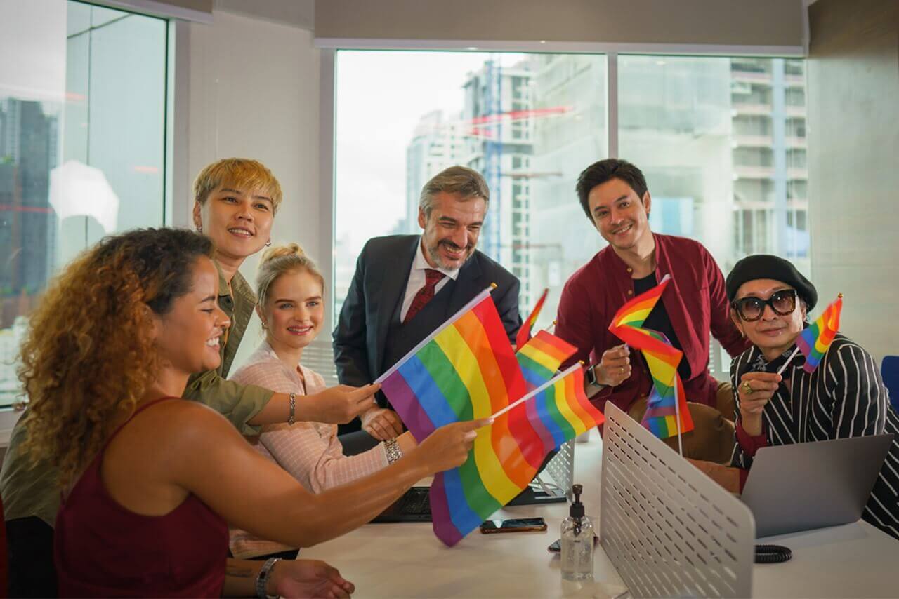 Pride at Work: Why Inclusion Still Matters in 2025 A diverse group of young people celebrating Pride outdoors, smiling and holding rainbow flags, symbolising LGBTQ+ inclusivity and community.