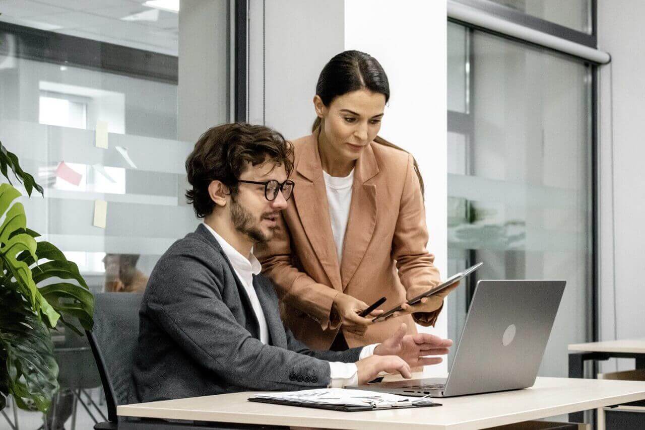 Two colleagues collaborating in a modern office, with a man seated at a desk working on a laptop and a woman standing beside him holding a tablet, discussing work