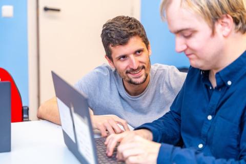 A friendly male assists a colleague in an office. The setting is bright and inclusive, with diverse employees engaging in activities.