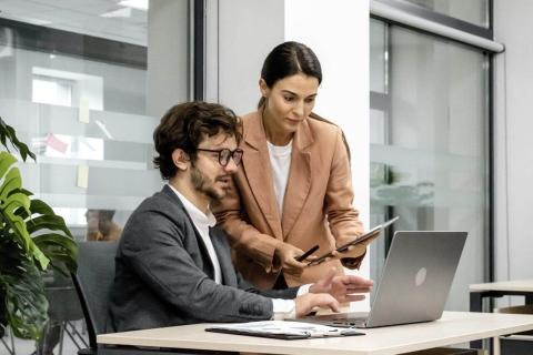 Two colleagues collaborating in a modern office, with a man seated at a desk working on a laptop and a woman standing beside him holding a tablet, discussing work