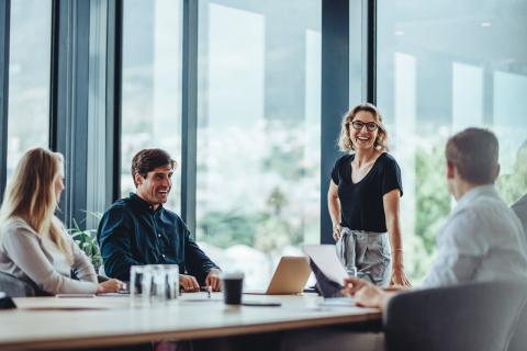 Two colleagues in a modern office lean in across a desk, engaged in a relaxed, friendly discussion, conveying trust, connection, and genuine workplace wellbeing.