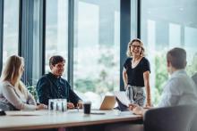 Two colleagues in a modern office lean in across a desk, engaged in a relaxed, friendly discussion, conveying trust, connection, and genuine workplace wellbeing.
